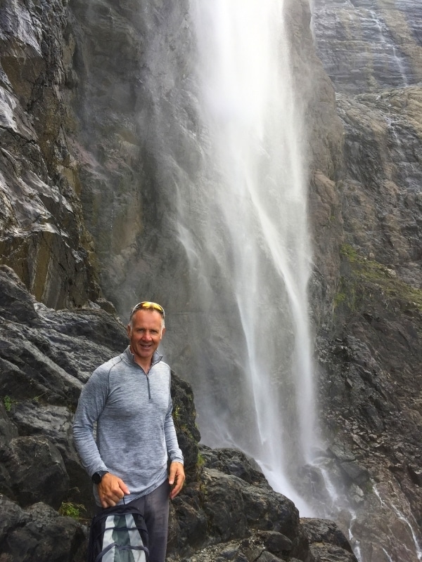 man standing under the spray from a tall waterfall