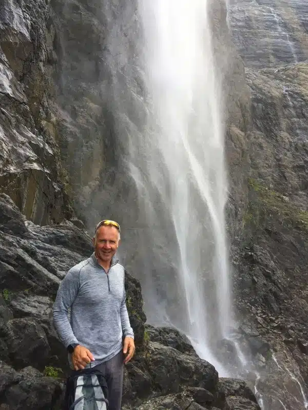 man standing under the spray from a tall waterfall