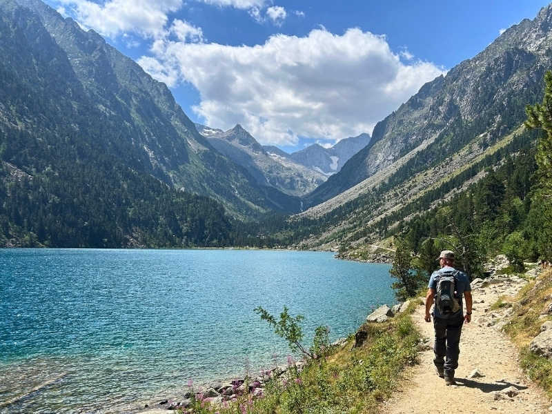 man in hiking gear walking along a rocky path by a turquoise alpine lake