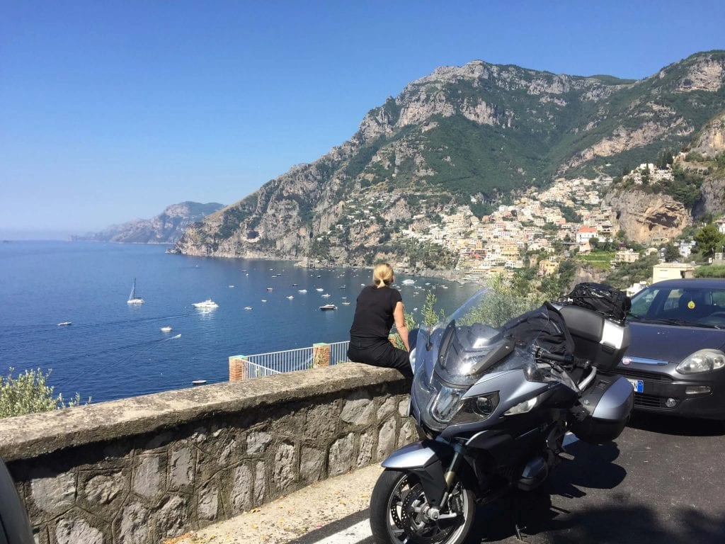 woman sitting on a wall looking at the sea view with a large motocycle parked nearby