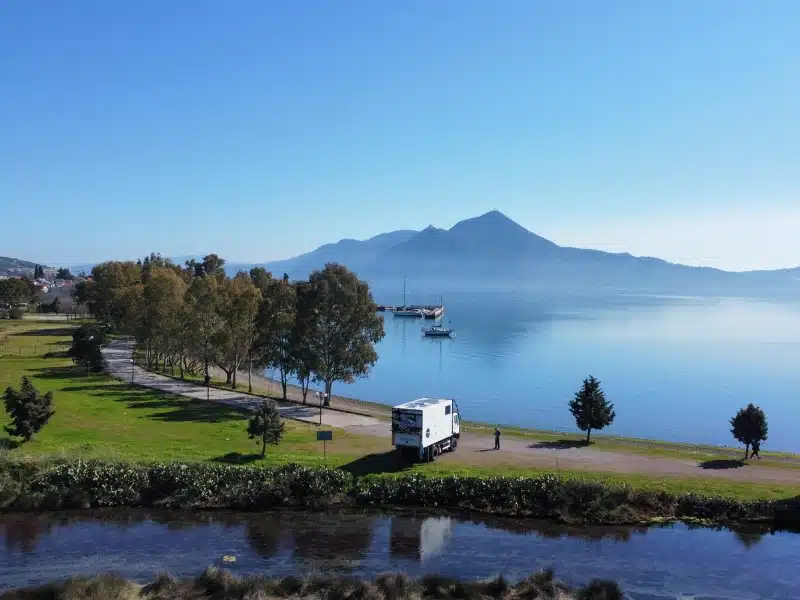 white overland truck parked bwteeen a clear river and blue sea with grass and trees in the picture