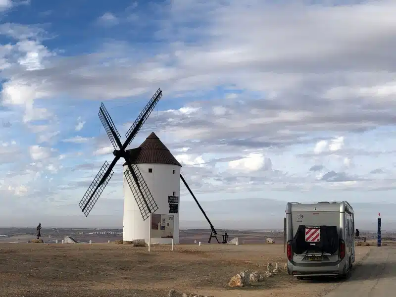 silver motorhome on a high point next to a white painted Spanish windmill with a conical roof and wooden sails