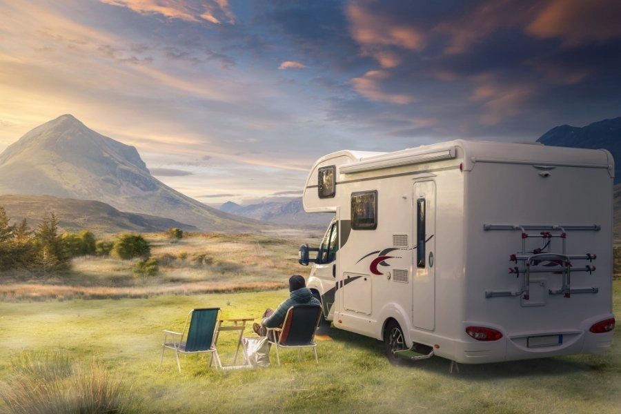 White motorhome parked in mountainsous area with a man sitting in a fold up camping chair outside