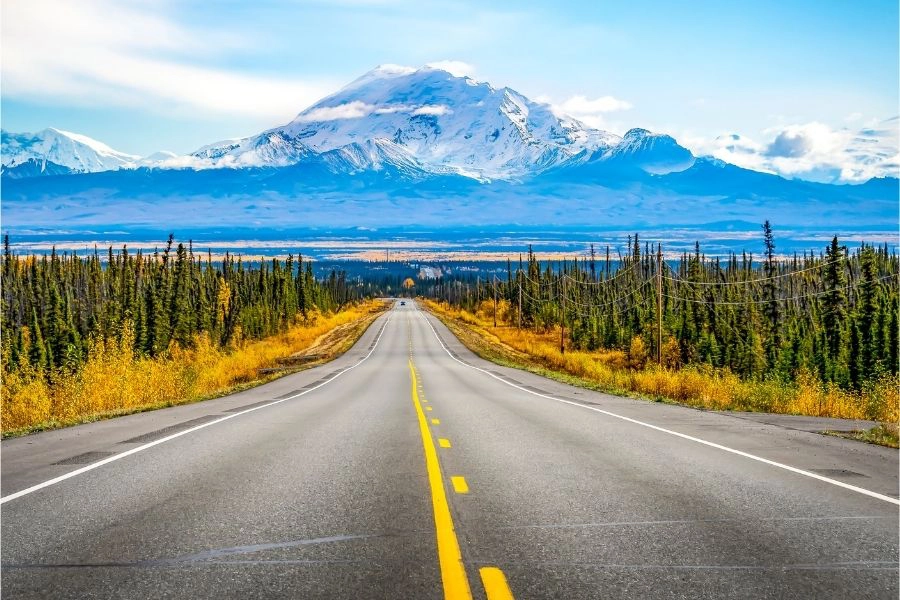 Ashphalt road with yellow markings going towards large mountains range with car in the far distance and trees along either side of the road.