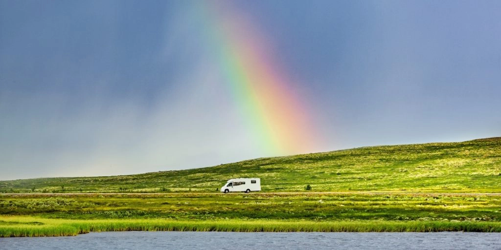 motorhome at the end of a rainbow on a road surrounded by grass with water in the foreground
