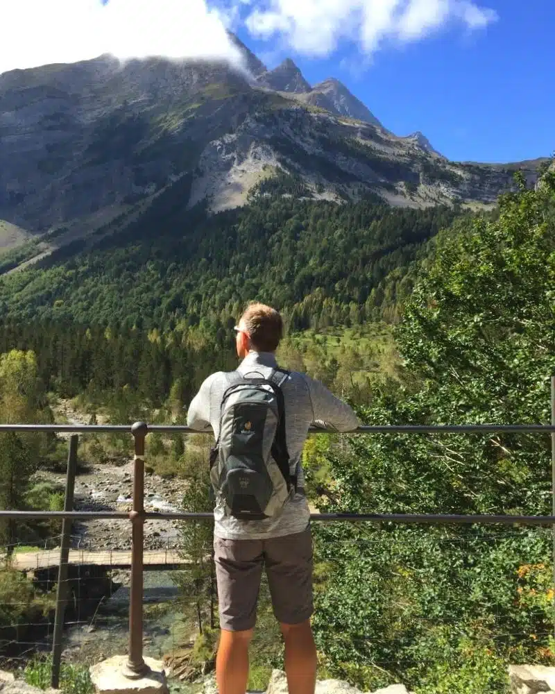 man in hiking gear carrying a rucksack standing on a bridge looking at high mountains