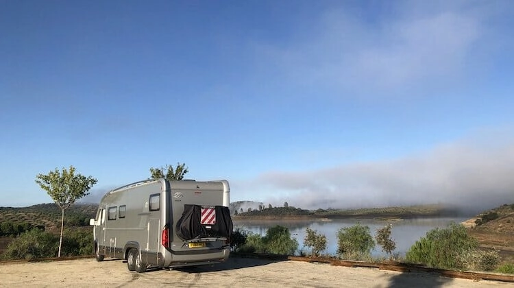 Large motorhome parked overlooking misty dam with small trees and blie skies