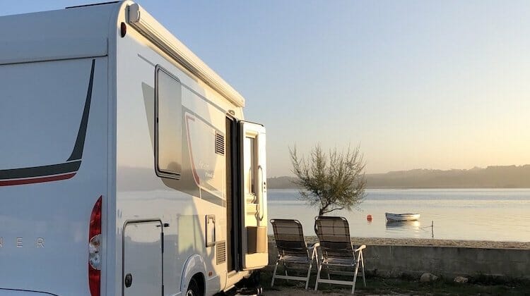 White motorhome over looking sea inlet with two camping chairs and small boat in the distance