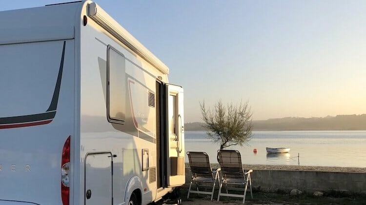 White motorhome over looking sea inlet with two camping chairs and small boat in the distance