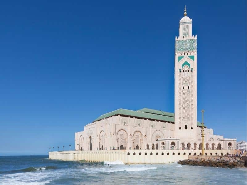 Large mosque with slender white tower, a green roof and decorative green tiling, surrounded by sea