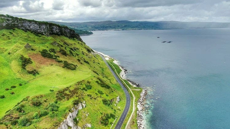 narrow road running by sea with green hills on the other side