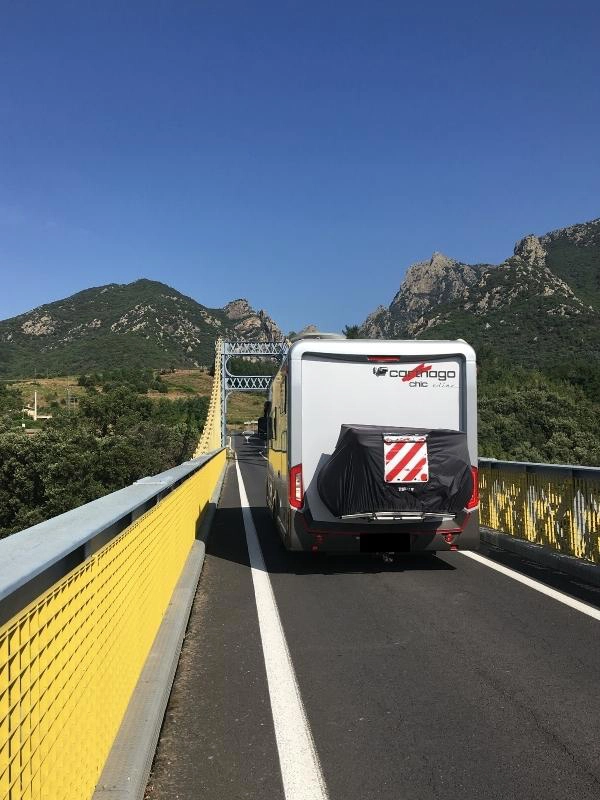 silver motorhome crossing a narrow bridge with yellow ironwork and small mountains in the background