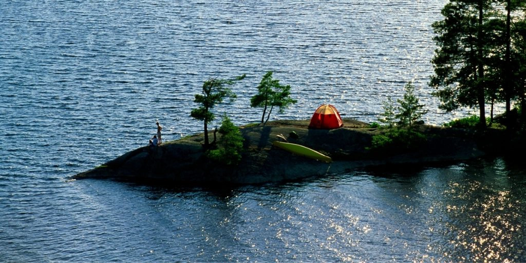 A small orange tent in a tiny peninsula with a few trees, a lime green kayak and two people looking at the surrounding water