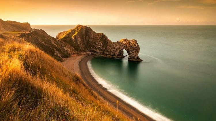 natural rock arch in the sea with a long sandy beacha dn grassy cliffs