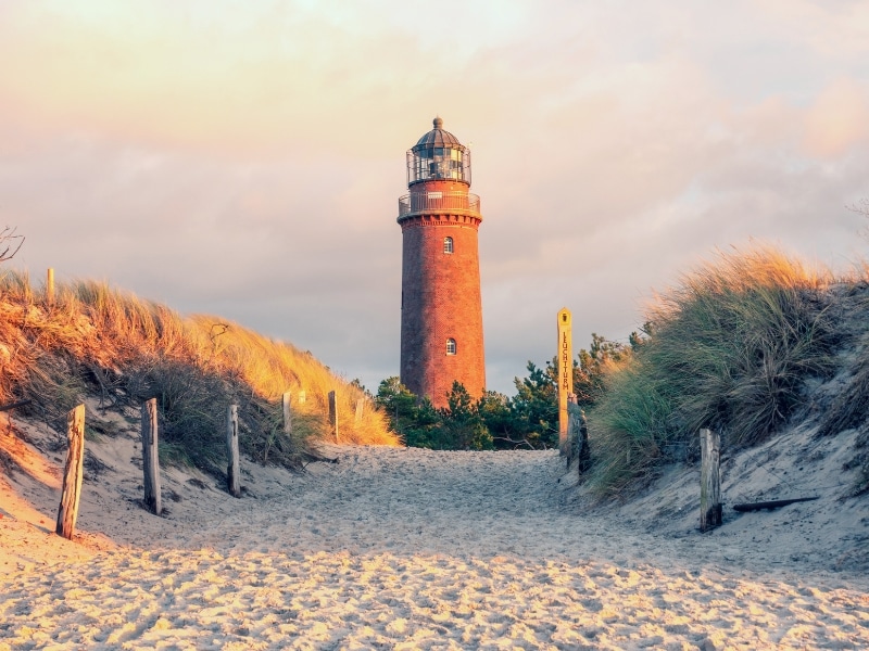 tall red brick lighthouse at the end of a sandy path lined with small grassy dunes