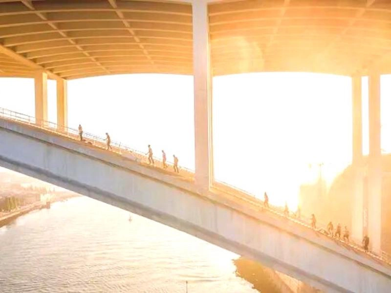 people climbing an arched bridge over a river with the sun rising behind them