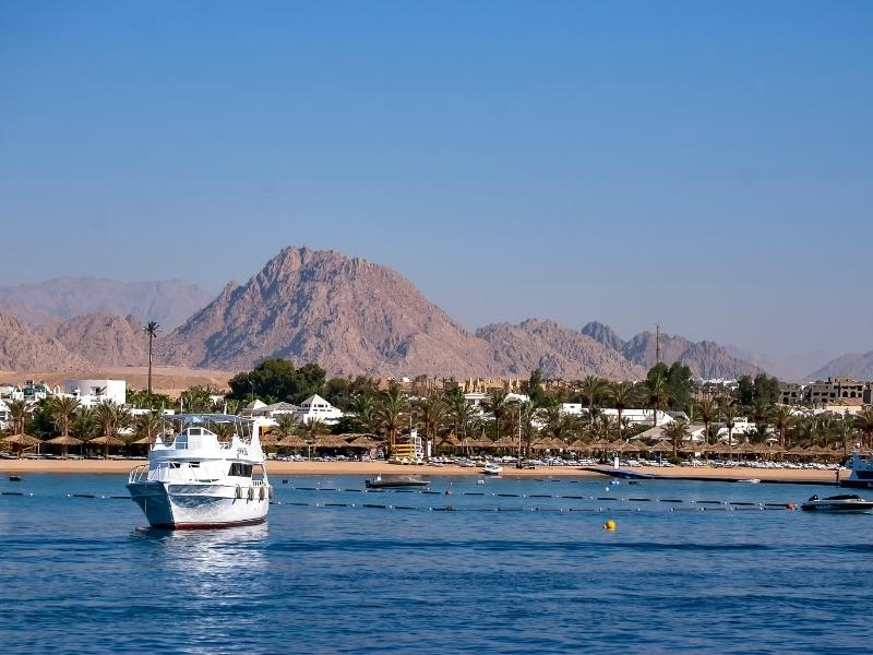 a white boat in a blue bay against rocky hills, palm trees ad low white buildings