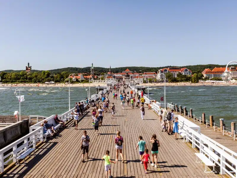 busy wooden pier leading to a beach and town of red roofed white houses