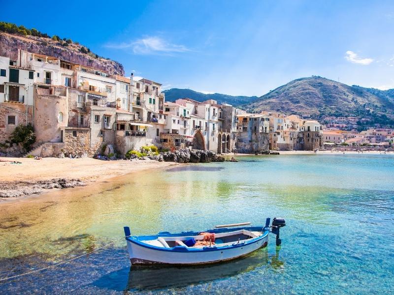 Small blue and white fishing boat in clear water by a beach and old houses.