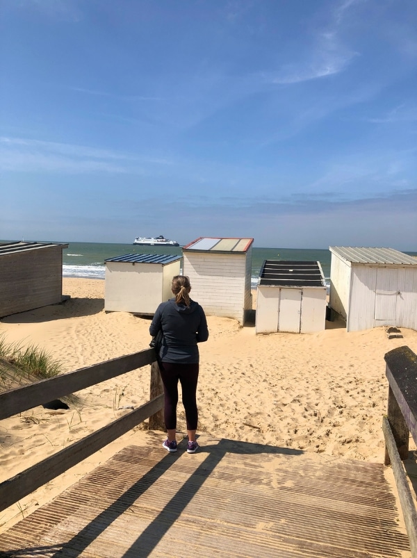 woman standing on the edge of a beach looking out to sea at a boat past beach huts