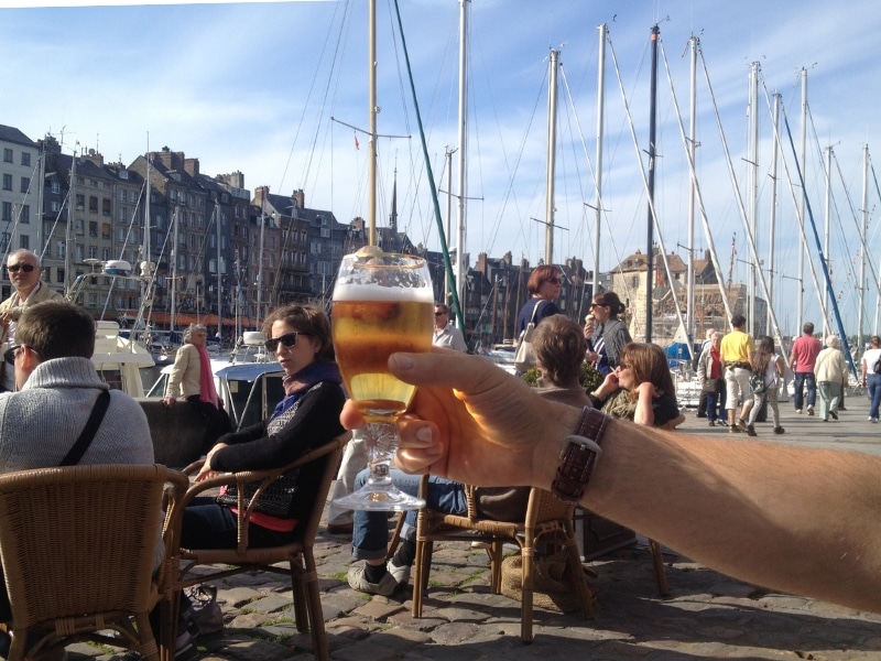 man holind g glass of beer aloft in front of masts and tall houses