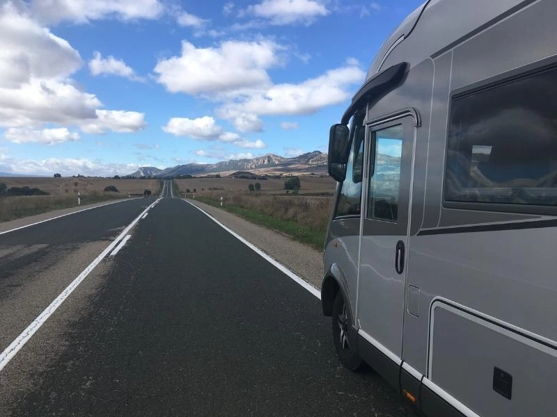Silver motorhome on an ashphalt road lined with fields, and mountains in the distance