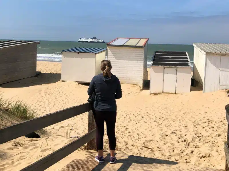 woman standing on a beach looking beyond beach huts to a ferry in the distance