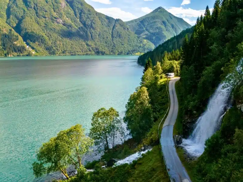 A winding road follows a fjord's edge, passing by a dramatic waterfall cascading down a rocky cliffside. Fjaerlandsfjorden, Fjord, Vestland, Norway