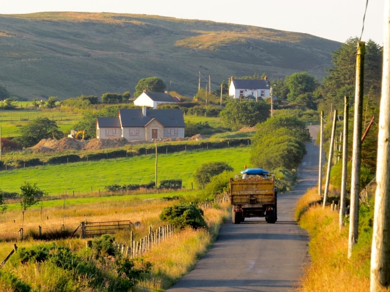 tractor moving along a rural road with a white farmhouse in the distance
