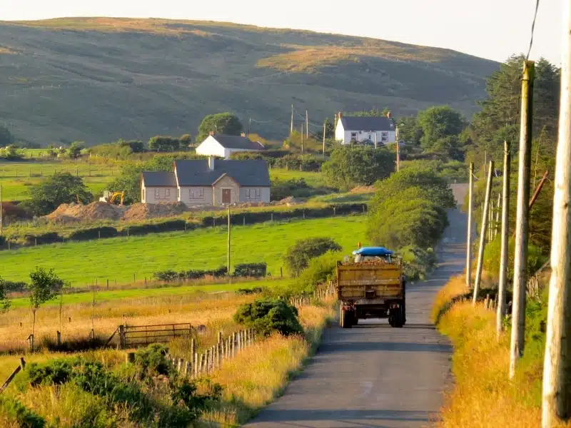 tractor moving along a rural road with a white farmhouse in the distance