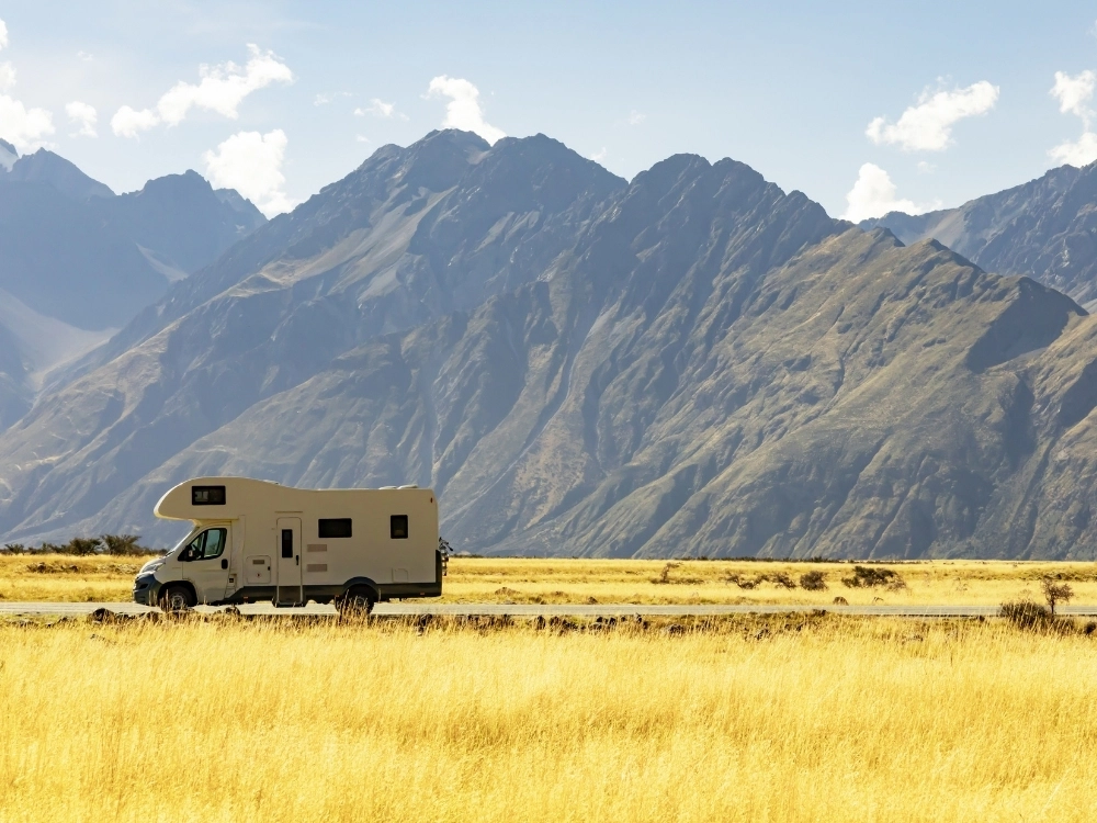 overcab motorhome driving on a road between yellow grass and mountains