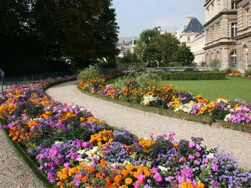 gravel path through colourful flower beds surrounded by grass, with historic buildings in the background