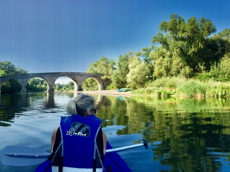 Woman in blue life jacket floating in a kayak with an arched bridge in the distance