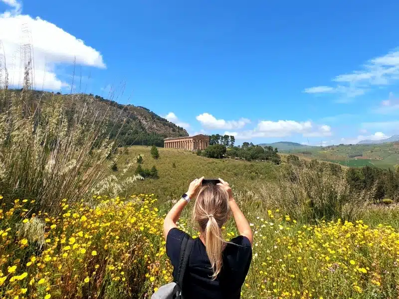 woman taking a photo of an ancient temple surrounded by grassy fields and yellow wildflowers