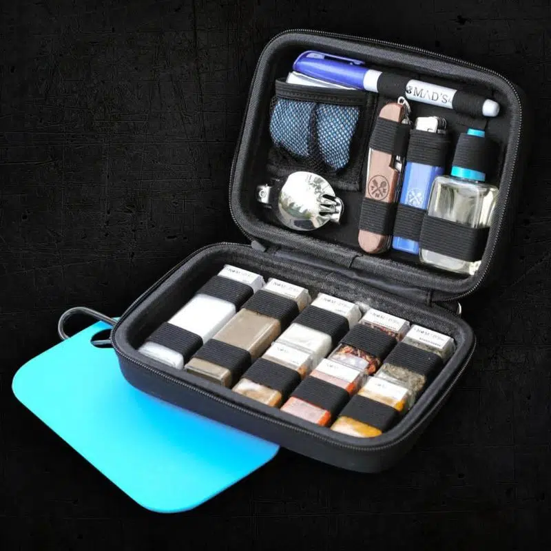 A small open case which included 8 small square jars of herbs & spices.  The other half of the case has a small bottle of oil, a lighter, penknife, spork and pen.  Hanging off the side on a carabiner is a small light blue chopping board.