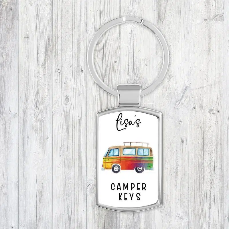 Thos shows a silver key ring with a rectangle with a rectangle fob which is white with a drawing of a multicoloured camper van.  Above the campervan the name Lisa is written in a cursive font, underneath the van is says Camper Keys.