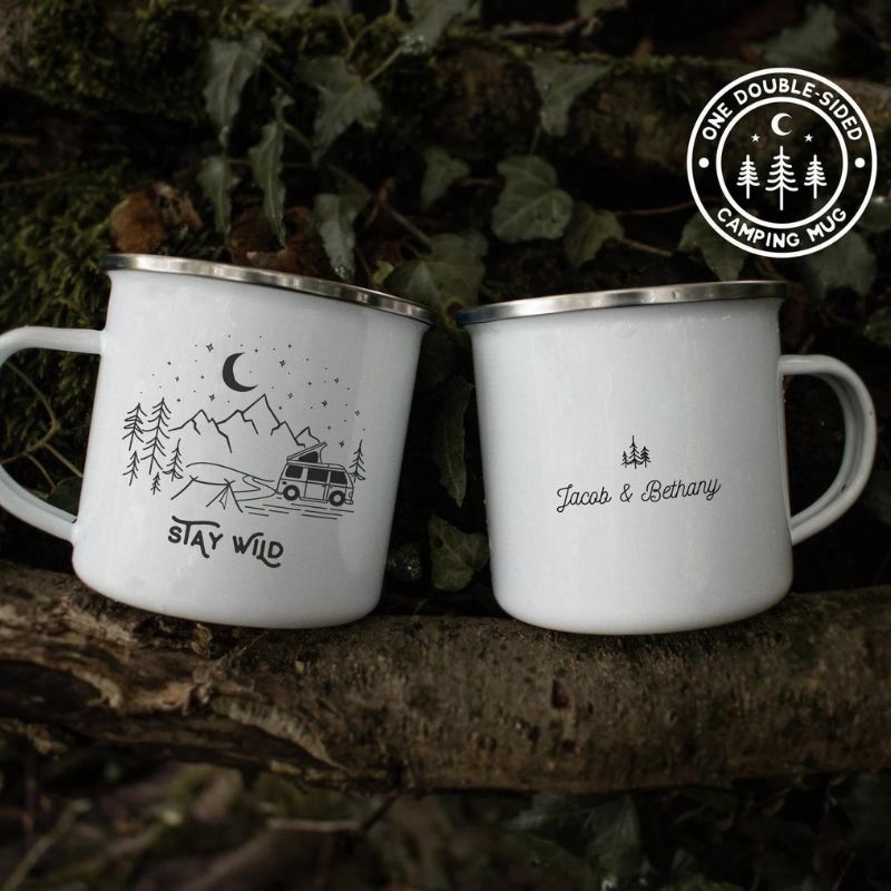 This image show 2 white enamel mugs with a silver rim.  One side of the mug has a drawing of a camper van in the mountains and the wording Stay Wild.  The other side has a drawings of three pine trees and the names Jacob& Bethany