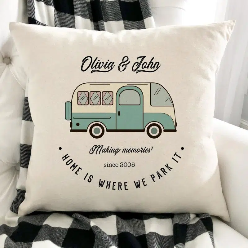 A cream pillow with a drawings of a retro campervan in teal, above ot has the names Olivia & John and below it says making memories, underneath that is sat since 2005 and then underneath that in an upside down semi circle is say HOME IS WHERE WE PARK IT.