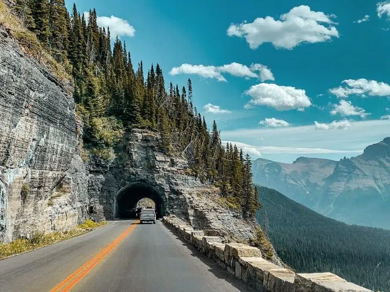 road with sheer cliffs to one side, going into a tunnel. There is a car on the road seen from the rear, and fir trees on top of the tunnel.