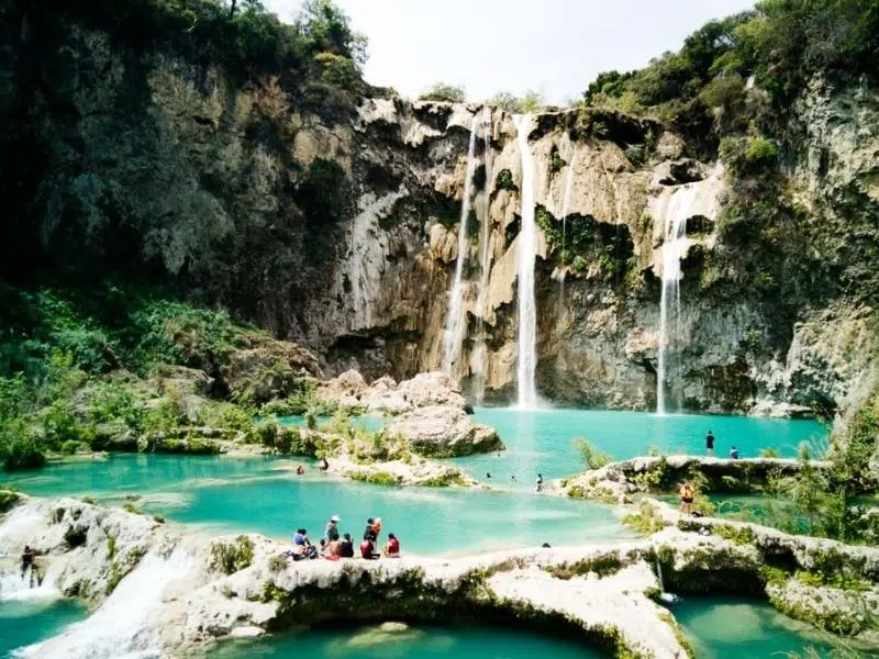 A waterfall with turquoise pools underneath and people taking photographs and swimming