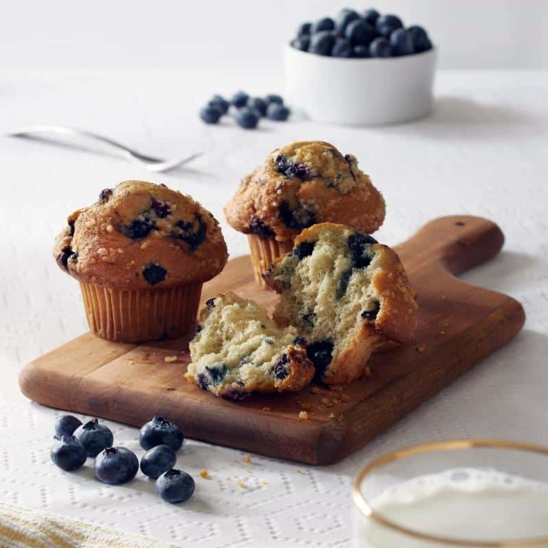 an image of three blueberry muffins on a small chopping board on a table, with loose blueberries.