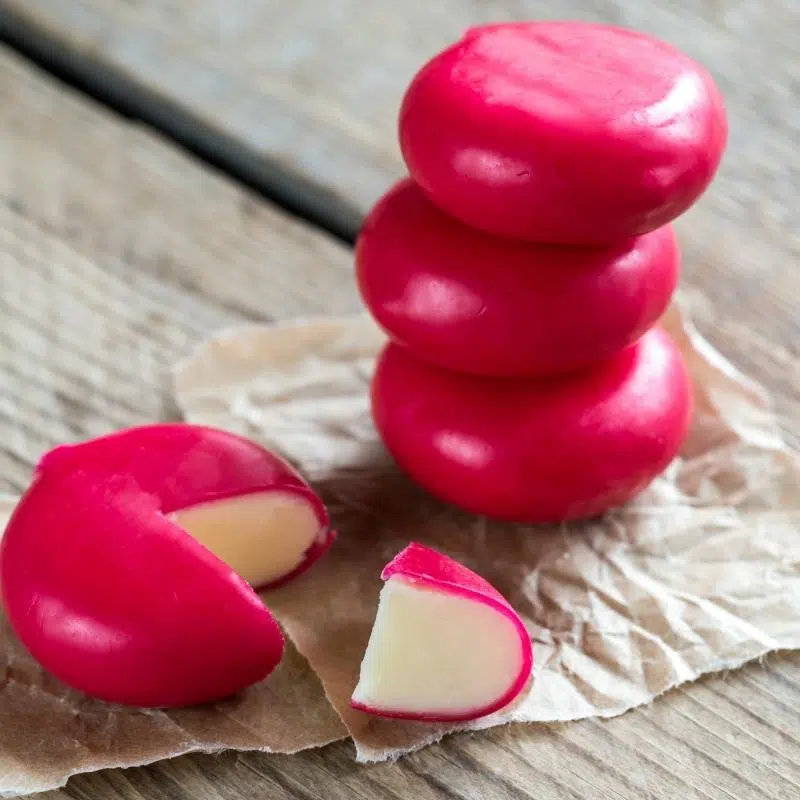 3 babybel cheeses in their red cases stacked, next to a babybel cheese with a slice cut out.