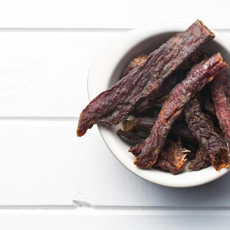 An image of a white table top, with a white bowl filled with beef jerky strips.