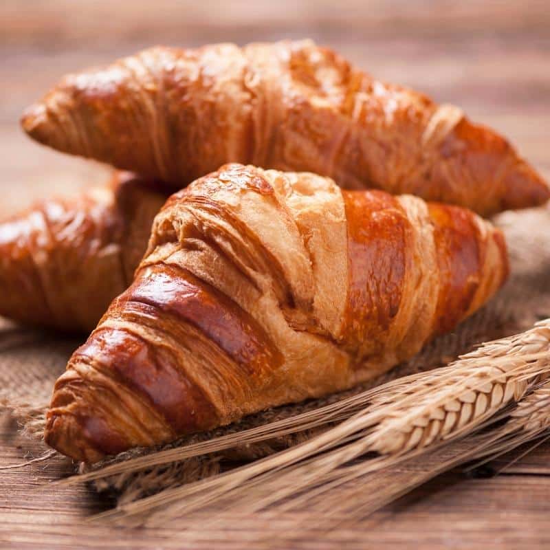 An image of three croissants on a wooden table with an ear of corn.