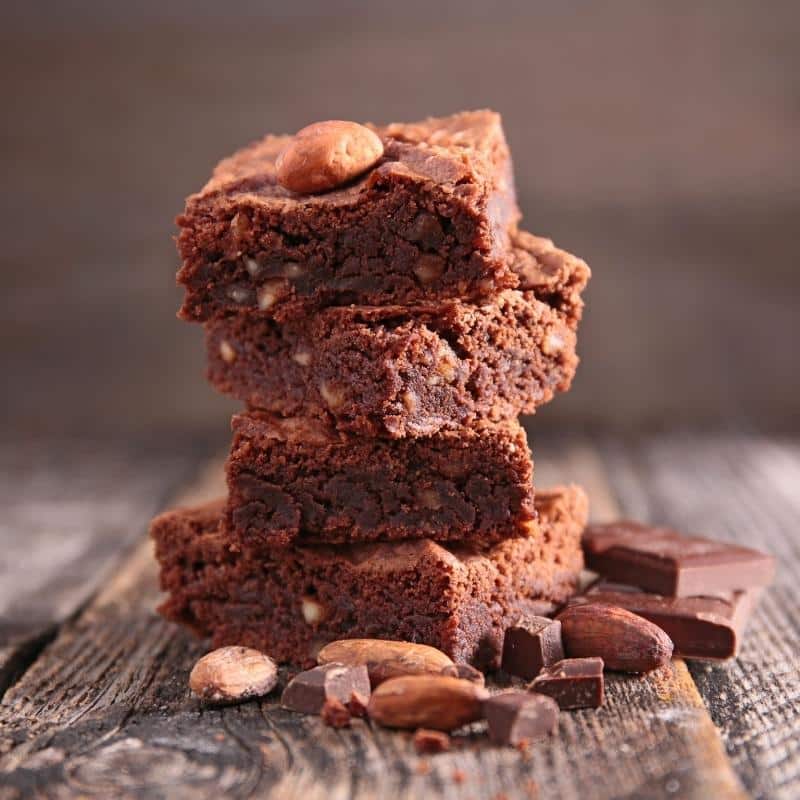 Am image of 4 chocolate brownies stacked on each other, on a wooden table with loose chocolate pieces and nuts.