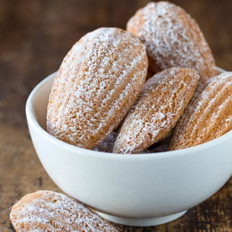 An image of a white bowl in a table filled with Madeleines dusted in icing sugar.