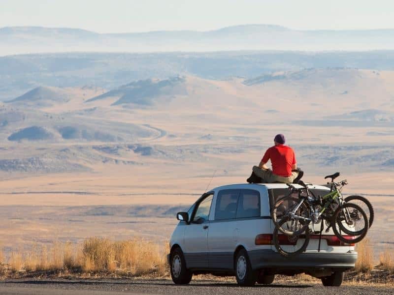 Man in a red tshirt sitting on a suv with bikes on a rack staring towards landscape of mountains and steppe