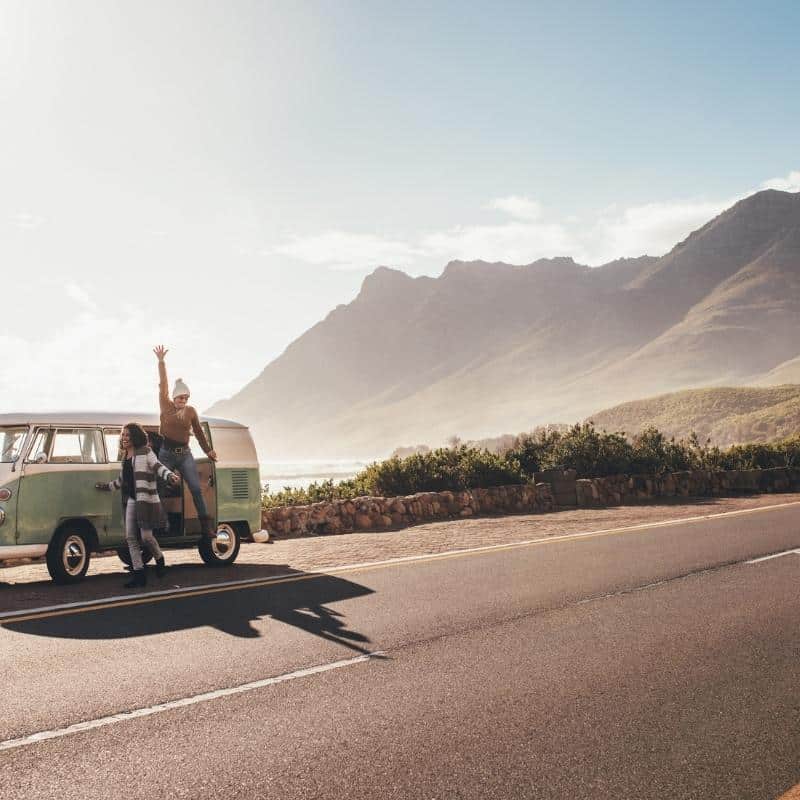 An image of a mint green VW Camper van pulled over on the side of the road, by the sea with mountains in the background.  Two wearing woolly hats are jumping out of the van, they're on a girls road trip.