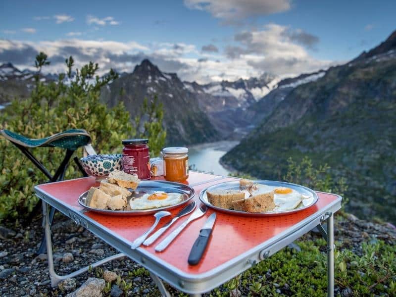 Folding camping table with two meals being served, in front of mountain and river view.