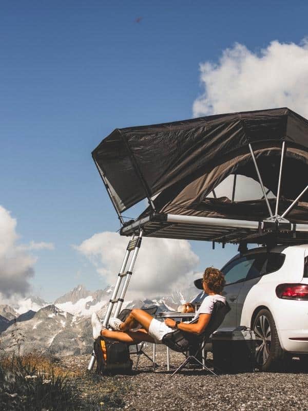 A white car with a large rooftop tent and a ladder for access. a Man sits in a camping chair looking at a view of te mountains.